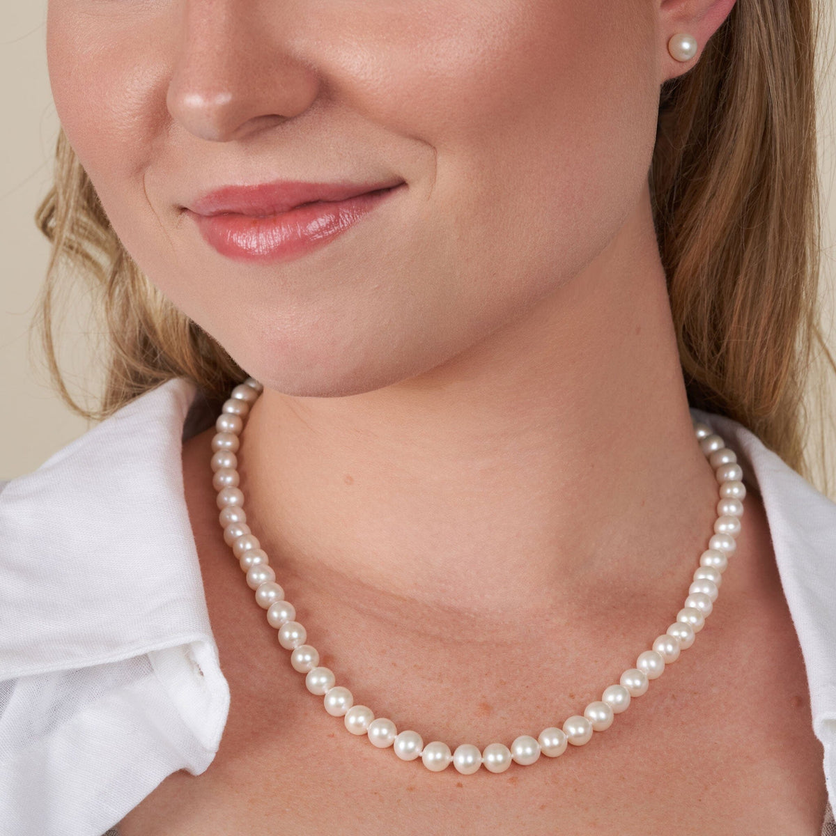 Close-up of a woman wearing a 6.5-7.0 mm AA+ White Freshwater Pearl Necklace and matching pearl stud earrings with a white blouse, highlighting the elegant pearl strand around her neck.