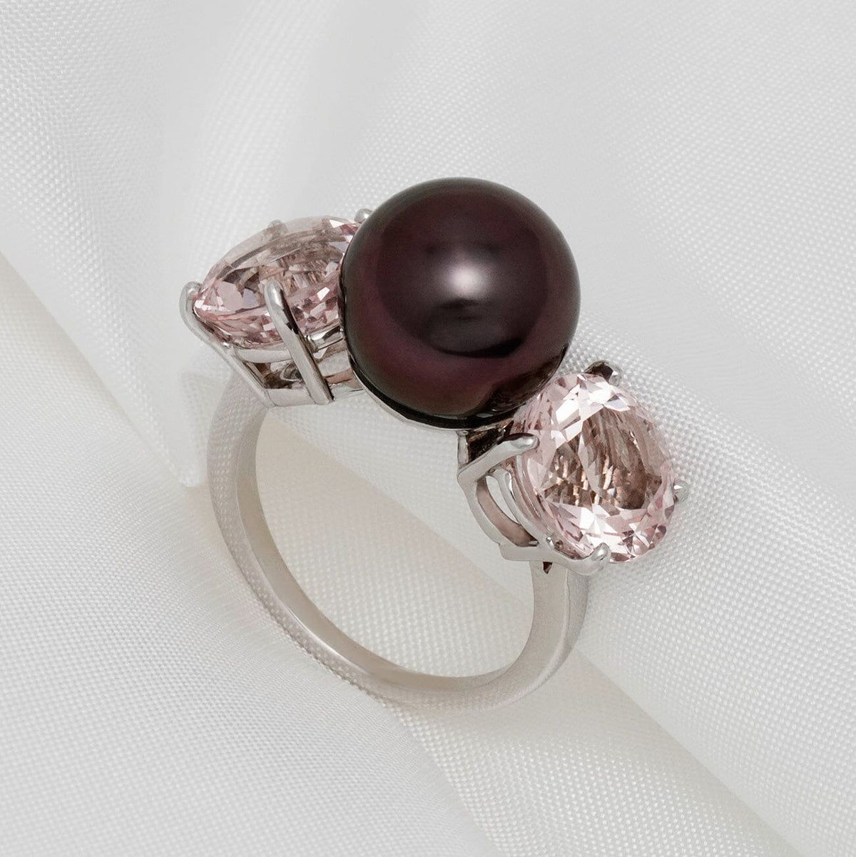 Assael Collection: 11.2 mm Tahitian Pearl and Morganite Ring with a dark lustrous pearl flanked by two oval pink morganite gemstones set on a sleek 14K white gold band against white fabric.