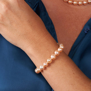 Close-up of a wrist wearing an 8.5-9.0 mm AAA Pink to Peach Freshwater Pearl Bracelet with round, lustrous pearls complementing a blue shirt.