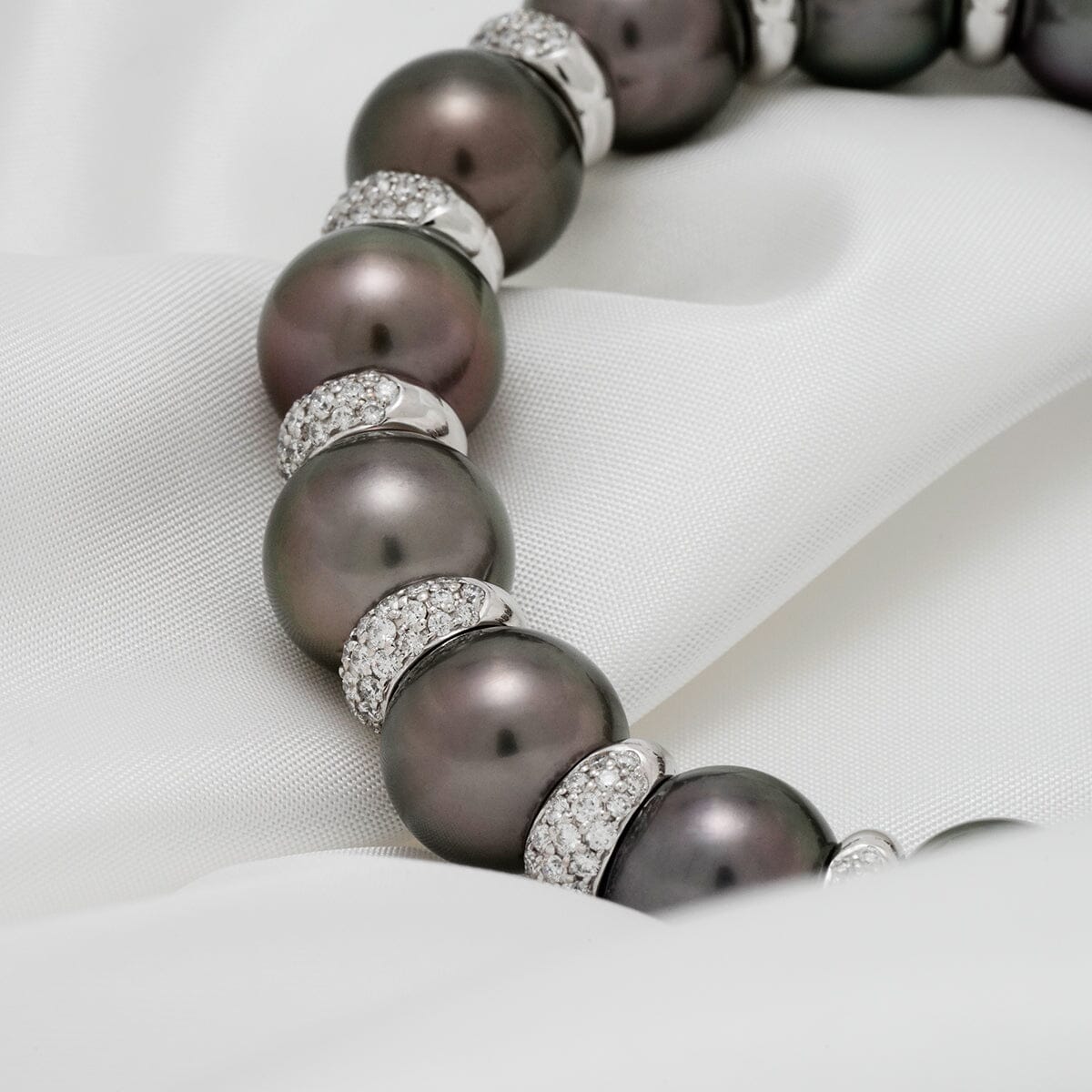 Close-up of Assael Collection: 11.0-12.7 mm Tahitian Pearl and Diamond Bracelet featuring lustrous dark pearls separated by sparkling diamond-encrusted spacers on a soft white fabric.