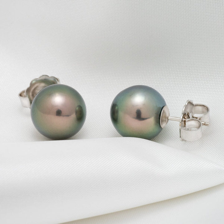 Assael Collection: 12.0–13.0 mm Tahitian Pearl Stud Earrings featuring lustrous round pearls with subtle green and lavender overtones set on silver posts against a white fabric background.