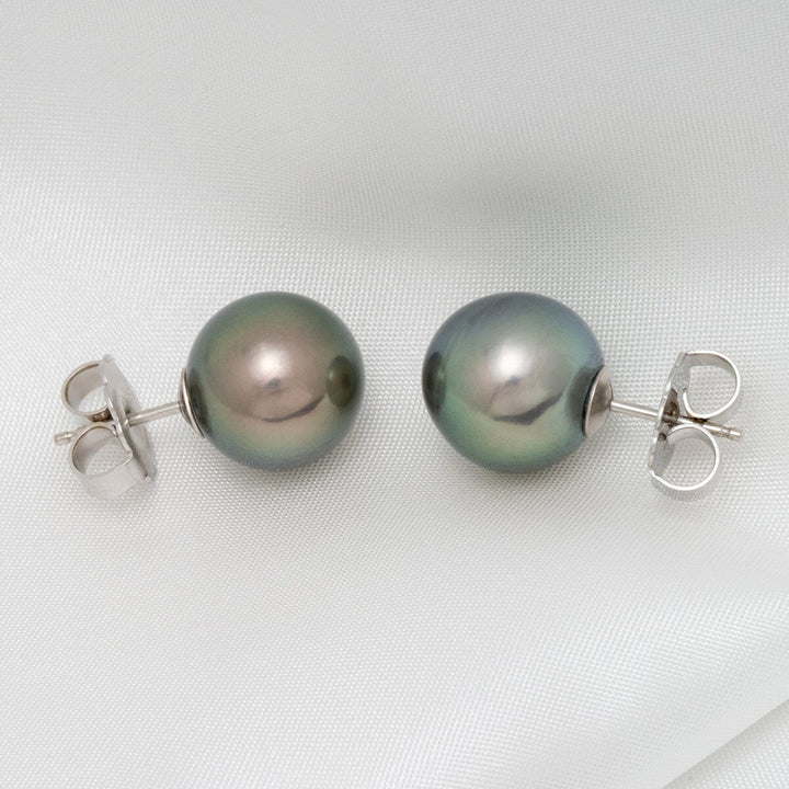 Assael Collection: 12.0–13.0 mm Tahitian Pearl Stud Earrings with lustrous dark pearls featuring subtle overtones, set on elegant silver posts on a smooth white fabric background.