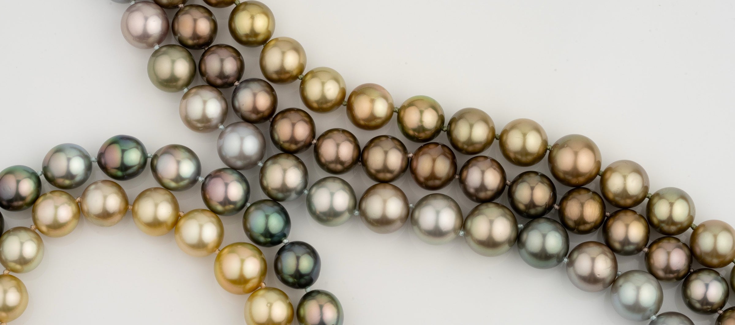 Close-up of multiple pearl strands featuring various shades including gold, bronze, green, and silver with a smooth, lustrous surface displayed against a plain background.