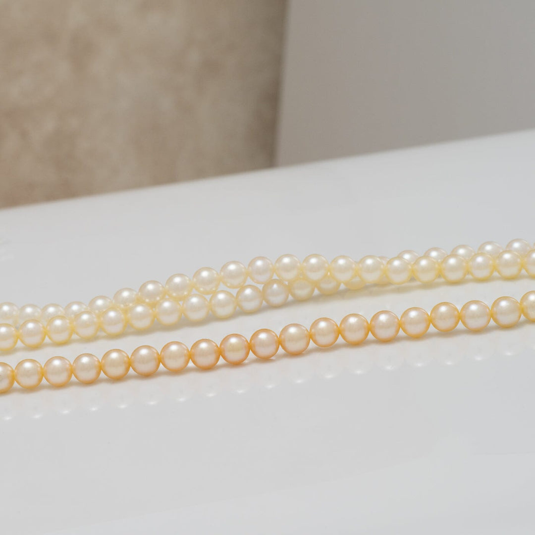 Two strands of TF-22: 4-4.5 mm Natural-color Round Akoya Ombré Pearl Strand showcasing smooth, lustrous pearls with a subtle transition of natural hues on a reflective surface.