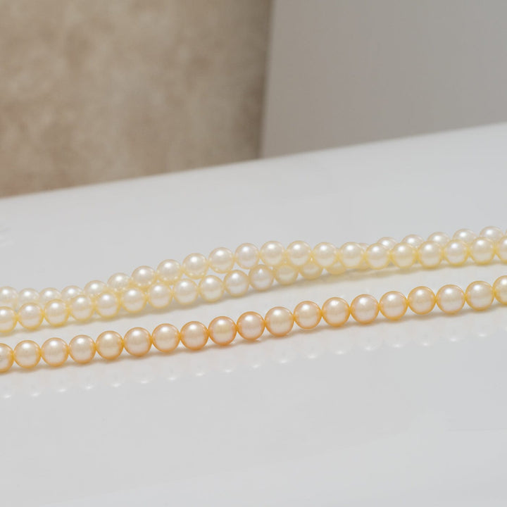 Two strands of TF-22: 4-4.5 mm Natural-color Round Akoya Ombré Pearl Strand showcasing smooth, lustrous pearls with a subtle transition of natural hues on a reflective surface.