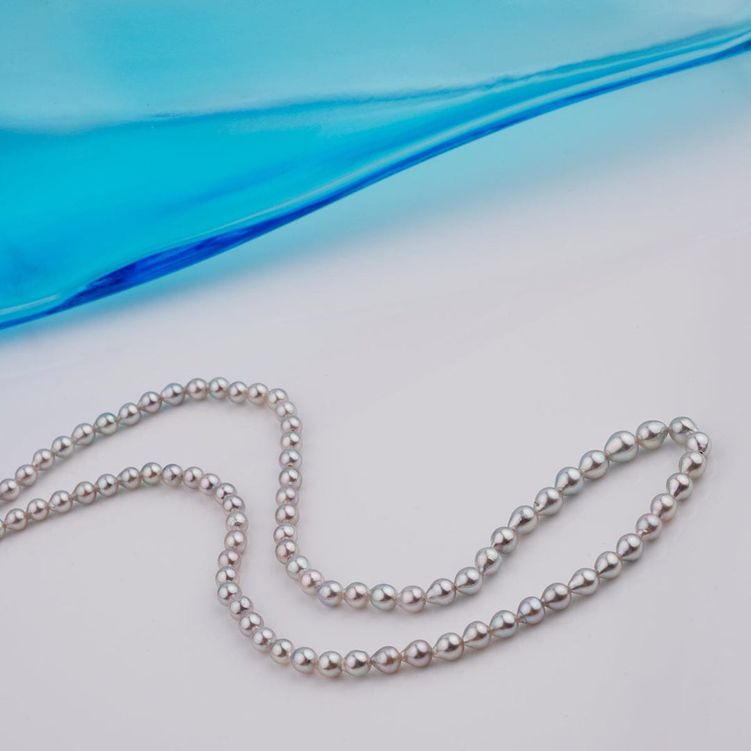 B-12: 3-4.5 mm Silver-blue Drop Akoya Strands, 16-inch Unfinished Strand elegantly displayed with lustrous cultured pearls softly reflecting light on a white surface near blue glass.