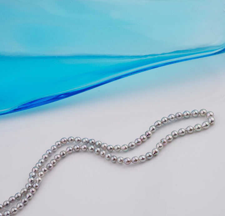 B-12: 3-4.5 mm Silver-blue Drop Akoya Strands, 16-inch Unfinished Strand displayed on a white surface with a smooth gradient blue glass element in the background.