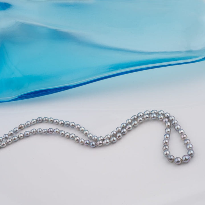 B-13: 3-4.5 mm Blue-silver Drop Akoya Strands, 16-inch Unfinished Strand displayed against a white background with a blue glass accent, showcasing fine cultured pearls.