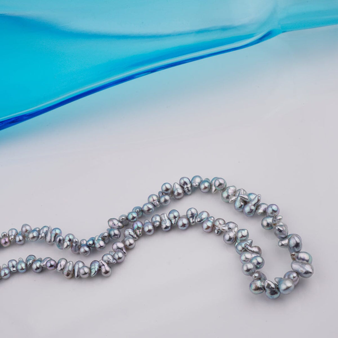 B-16: 2.5-4 mm Top-drilled Blue Baroque Akoya Strands, 18-inch Unfinished Strand with irregularly shaped blue akoya pearls displayed on a light surface next to a blue glass object.