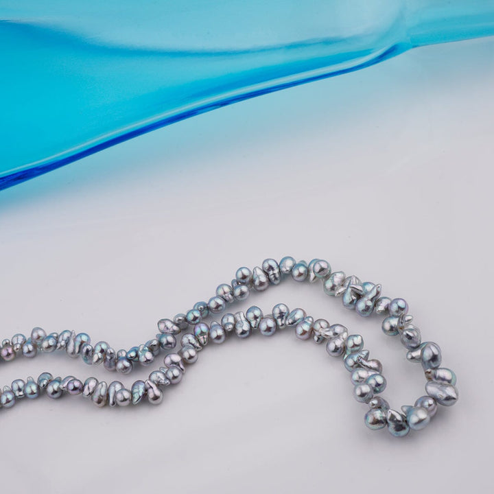 B-16: 2.5-4 mm Top-drilled Blue Baroque Akoya Strands, 18-inch Unfinished Strand with irregularly shaped blue akoya pearls displayed on a light surface next to a blue glass object.