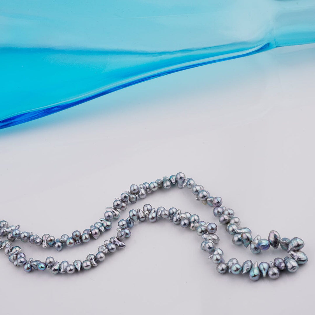 B-16: 2.5-4 mm Top-drilled Blue Baroque Akoya Strands, 18-inch Unfinished Strand displayed on a light surface with a blue glass accent, showcasing unique irregular pearls for jewelry making.