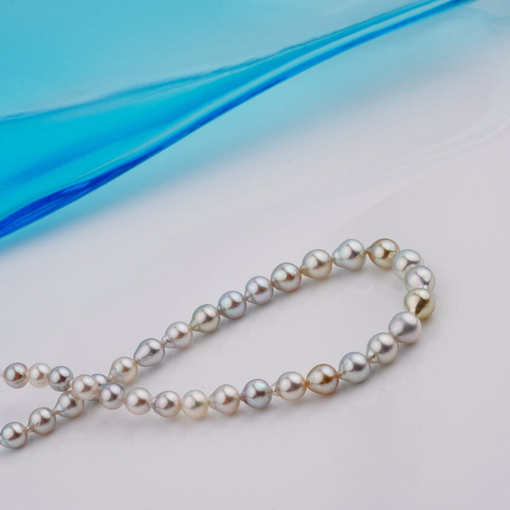 B-18: 4.5-6 mm Multicolor Drop Akoya Pearl Bracelets, 7.25" Unstrung displayed with lustrous pearls in various soft hues on a smooth surface with a blue glass accent in the background.