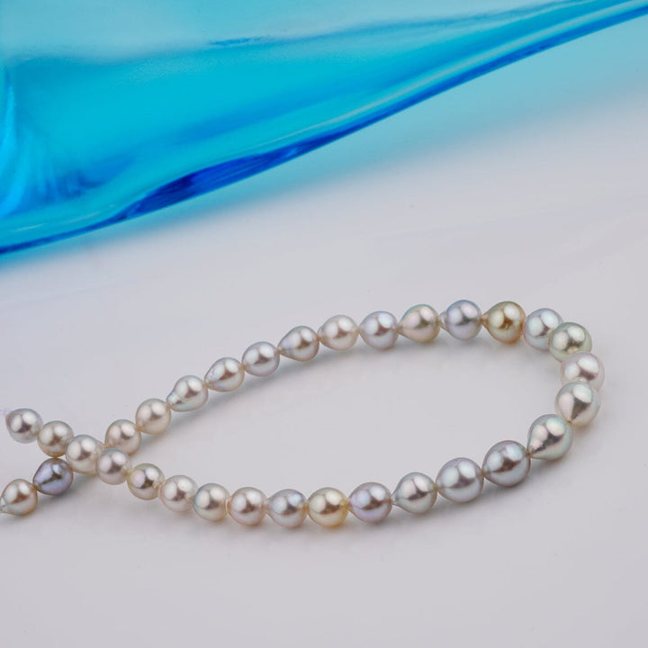 B-18: 4.5-6 mm Multicolor Drop Akoya Pearl Bracelets, 7.25" Unstrung displayed on a white surface with a blue glass accent, showcasing varied natural pearl tones and drop shapes.